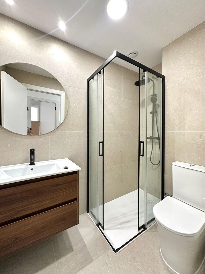 Baño reformado en Vigo con plato de ducha, mampara de perfil negro, mueble suspendido de madera y espejo redondo